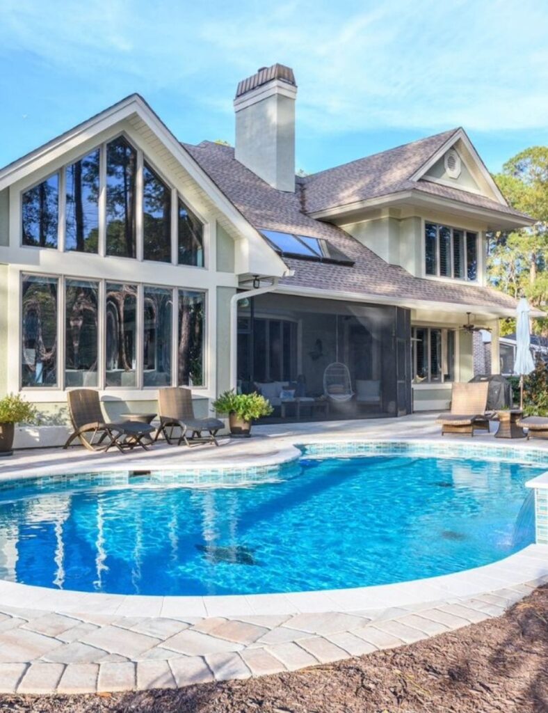 a curving in-ground pool with clean tile accents and a spacious stone deck perfect for lounging. Framed by a beautiful home with floor-to-ceiling windows and cozy outdoor seating, it’s the ideal spot to unwind, entertain, and enjoy serene poolside living