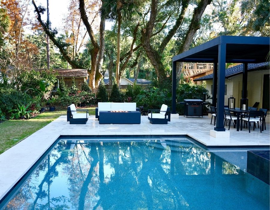 a modern, serene backyard with a geometric pool framed by light stone decking. The space features a stylish black-and-white outdoor lounge set and a shaded pergola covering a dining and grilling area, perfect for entertaining. Surrounded by lush tropical greenery and mature trees, the setup blends contemporary design with natural tranquility