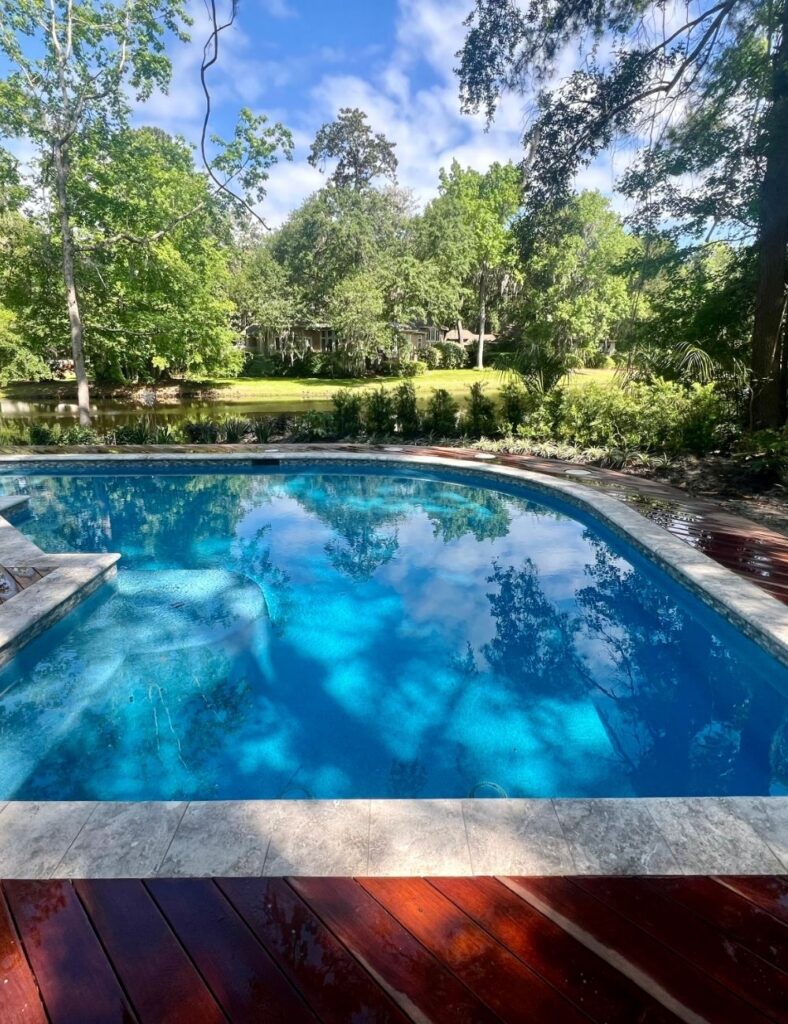 This backyard oasis blends natural serenity with elegant design. The curved-edge pool is framed by rich, reddish-brown wooden decking and light stone coping, creating a striking contrast. Reflections of the sky and surrounding trees ripple across the brilliant blue water, while lush greenery and a tranquil pond beyond add to the peaceful, secluded atmosphere—perfect for a relaxing escape right at home