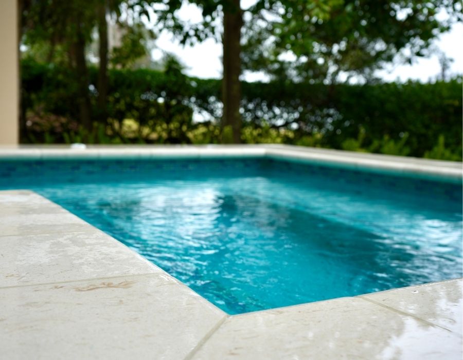 backyard pool framed by creamy stone tiles, with glistening blue water reflecting the soft, natural light and lush greenery providing a private, tranquil escape