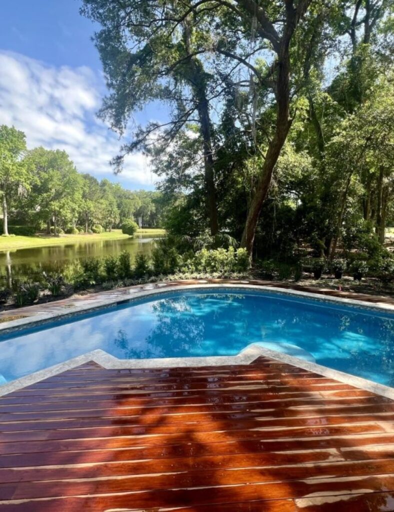 Backyard swimming pool with a wooden deck overlooking a tranquil pond and lush green trees on a sunny day