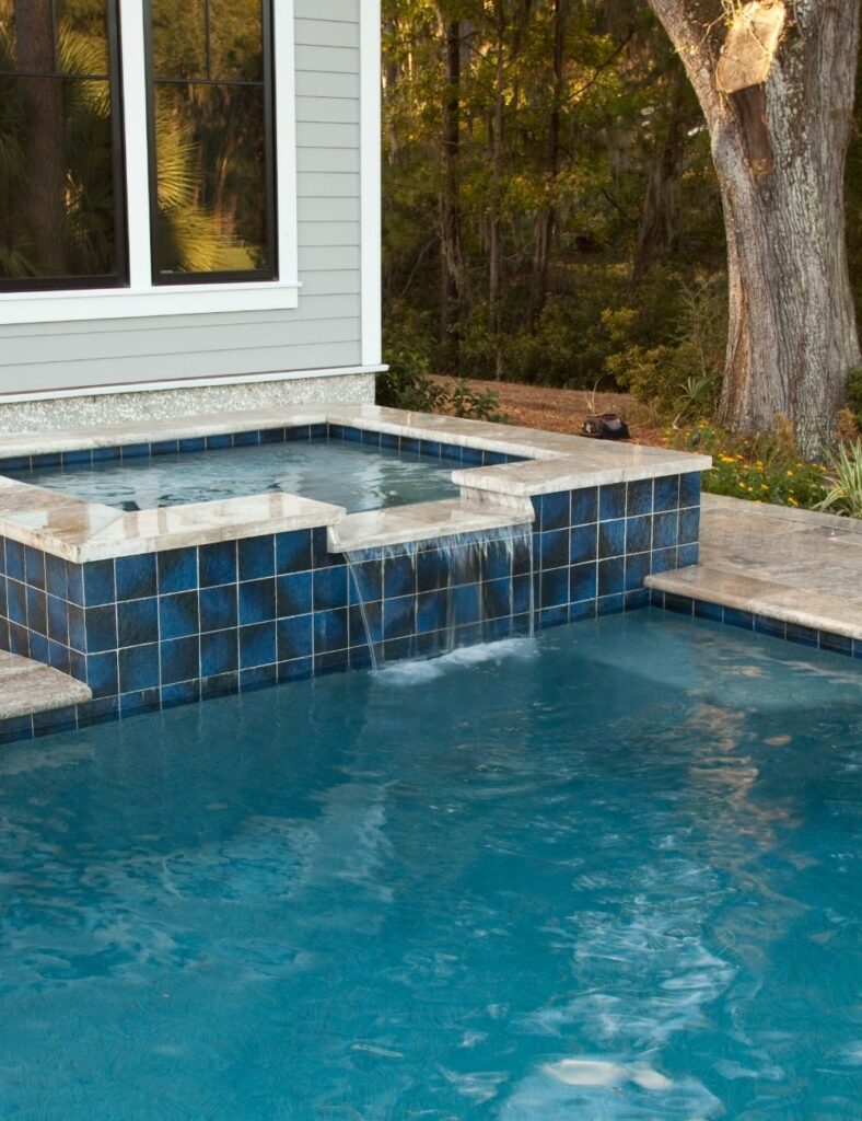 pool features a raised spa with a spillover waterfall into the main swimming area, surrounded by natural stone tile decking and tranquil wooded views—creating a serene retreat just steps from the home