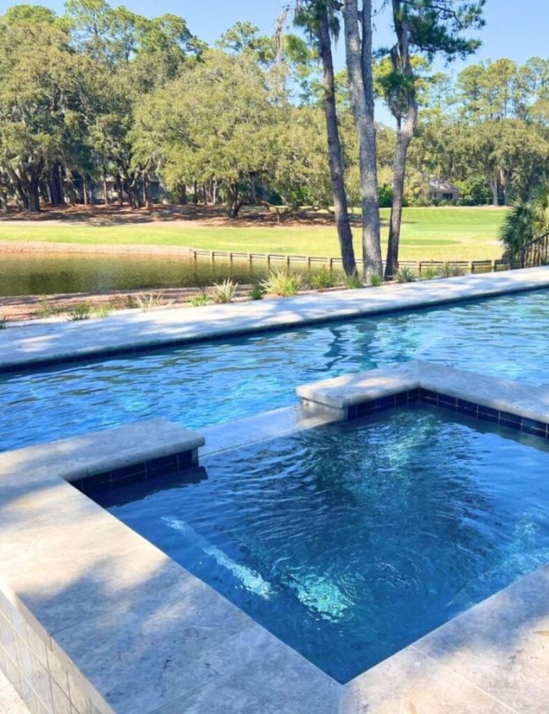 elevated pool and spa combo with a seamless edge overlooking a peaceful golf course and tranquil pond. The modern stone surround enhances the elegant design, while the clear blue water reflects the surrounding trees and sky. It's a perfect blend of luxury and natural beauty—ideal for relaxing with a view