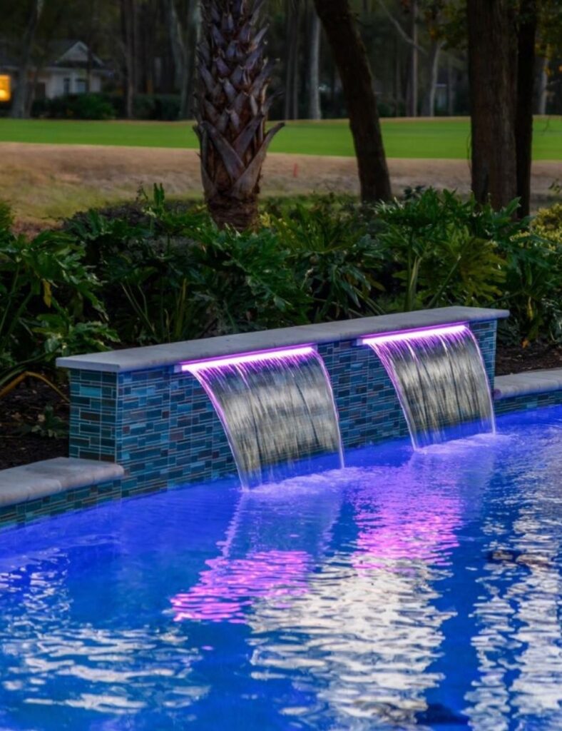 a modern pool with dual illuminated waterfalls cascading from a sleek, tiled wall, enhanced by stunning purple LED lights that reflect beautifully across the evening water