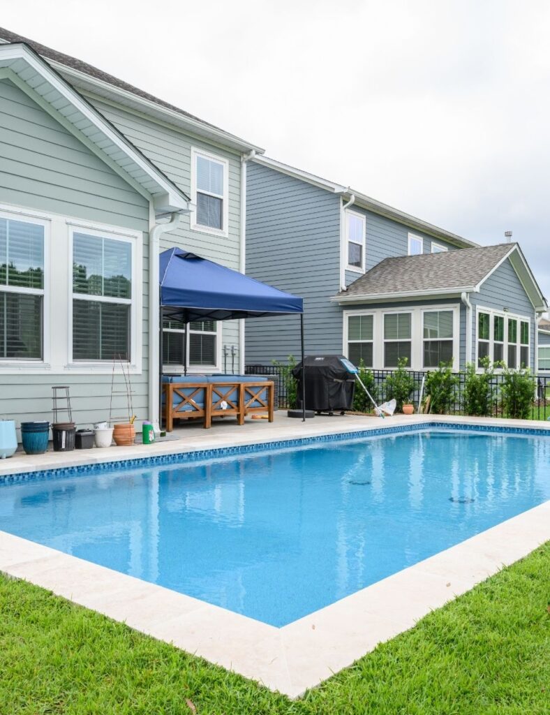 rectangular backyard pool bordered by crisp white coping and lush green grass. Set beside a two-story home with soft sage siding and white-trimmed windows, the pool area includes a shaded seating nook with a blue canopy and a few potted plants, creating a charming and functional family-friendly space perfect for relaxing or entertaining