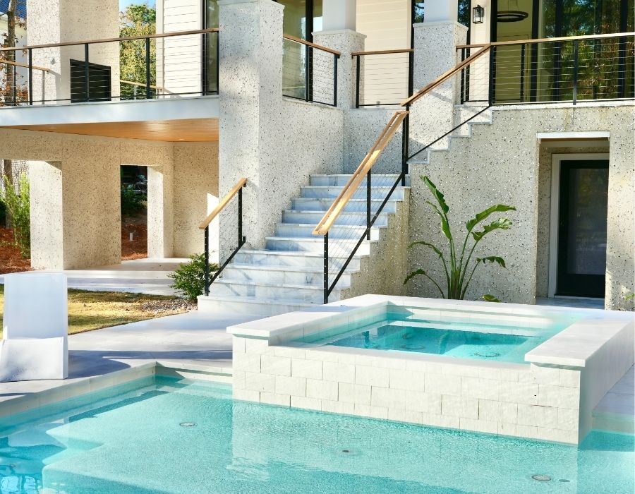 This stunning poolscape pairs crisp modern lines with bright, serene aesthetics. The elevated spa features a clean, geometric design with light stone facing and spills into a refreshing turquoise pool below. Surrounded by sleek white pavers and lush tropical accents, the space is complemented by a grand staircase and stylish railings, creating a luxurious and contemporary backyard retreat
